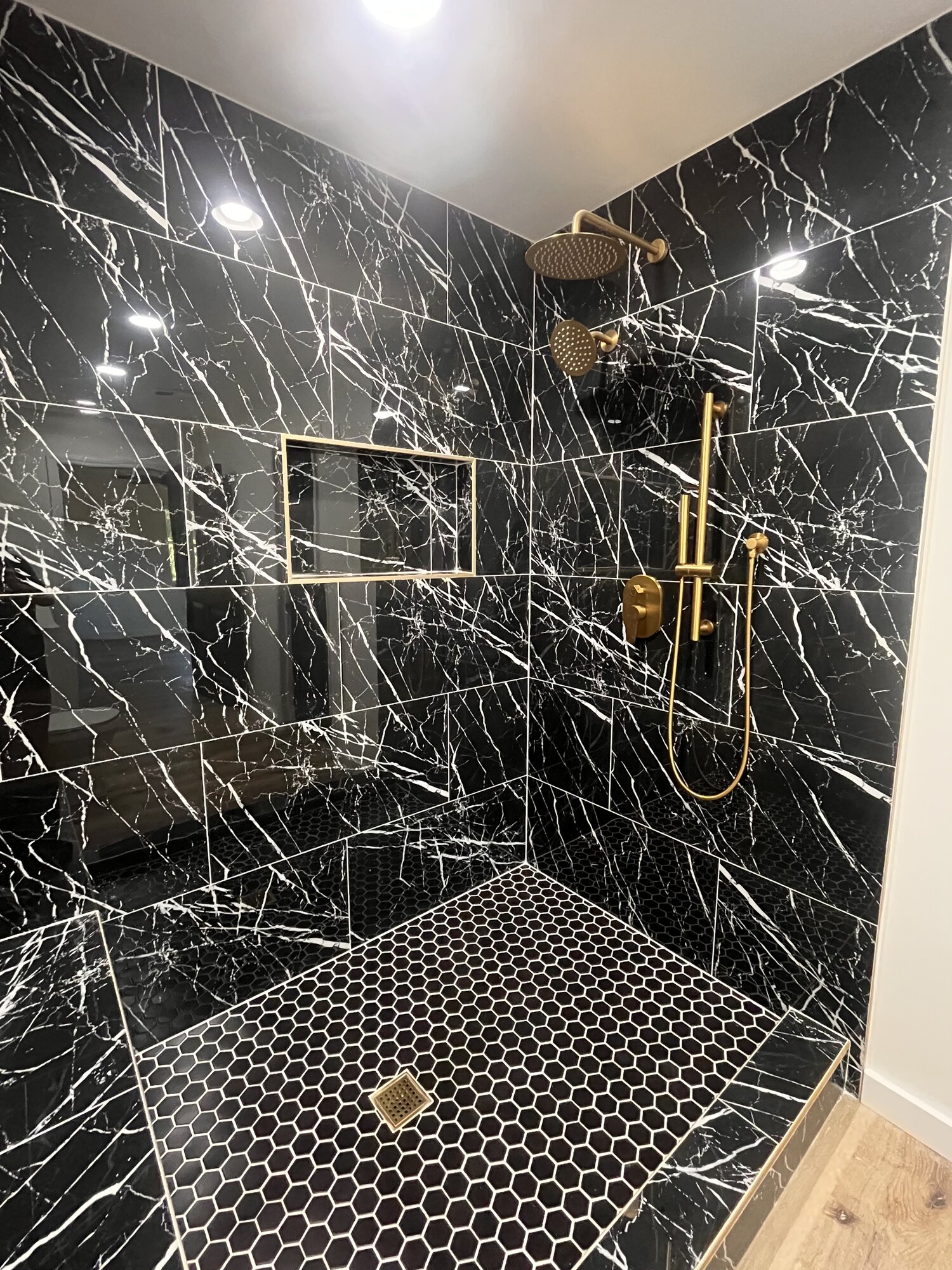 Dramatic black marble walk-in shower with brushed brass rain head, hand shower, and black hex mosaic floor